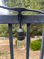 Drop-style fence clip holding a lit Edison-style string light bulb on a metal railing — Groovy Lanterns