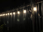 Drop-style fence clips holding a row of lit string light bulbs along a fence at night — Groovy Lanterns